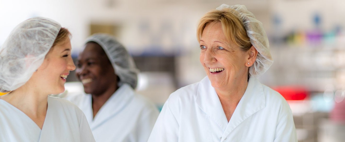 Zwei Mitarbeiterinnen mit Haarnetz und weißer Arbeitskleidung unterhalten sich und lachen in der Klinikküche.
