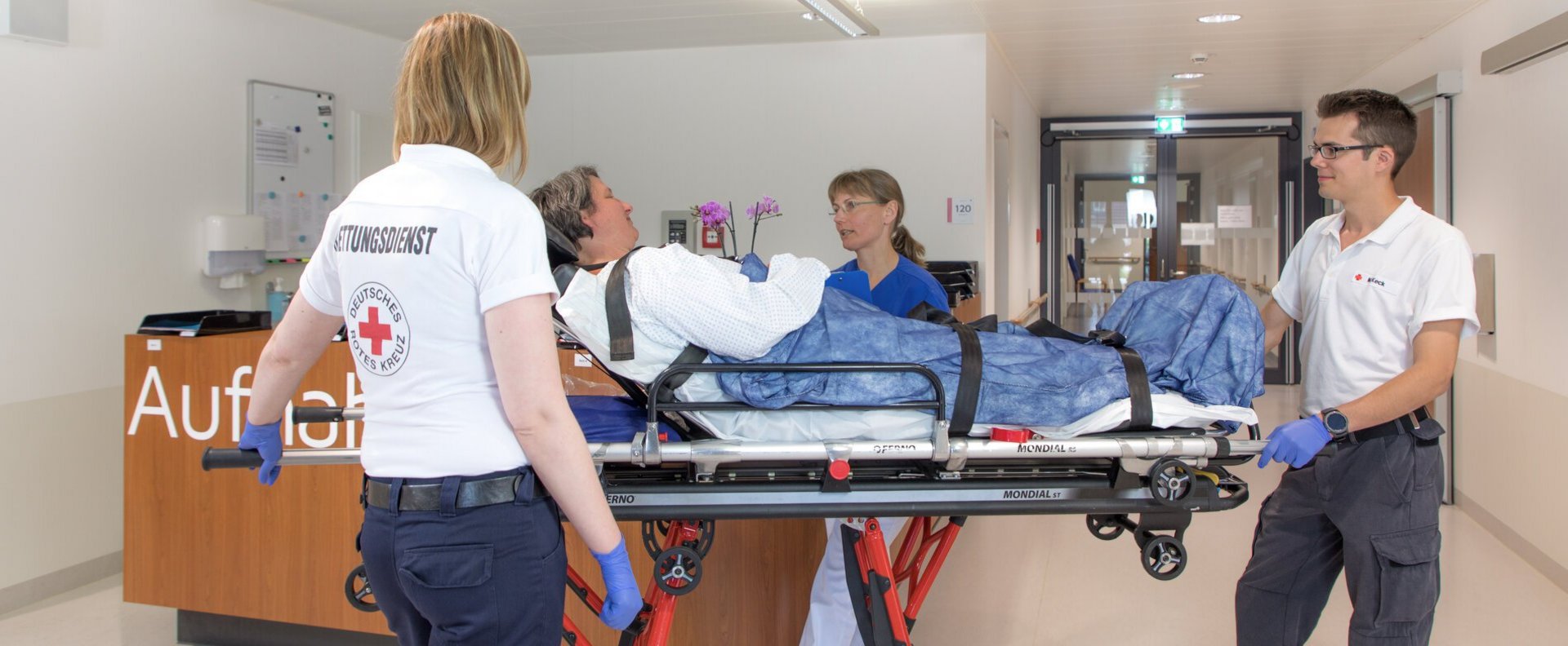 Patientin wird auf fahrbarer Liege von zwei Rettungsassistenten auf die Aufnahmestation gebracht und von Pflegefachfrau in Empfang genommen.