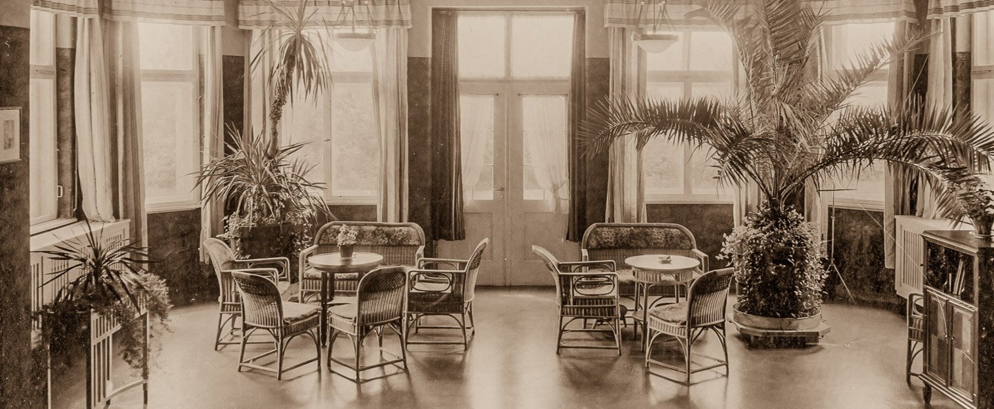 Historisches Bild vom Gartensaal des Genesungsheims für Tropenheimkehrer mit einer großen Fensterfront, Sitzgelegenheiten und tropischen Pflanzen.