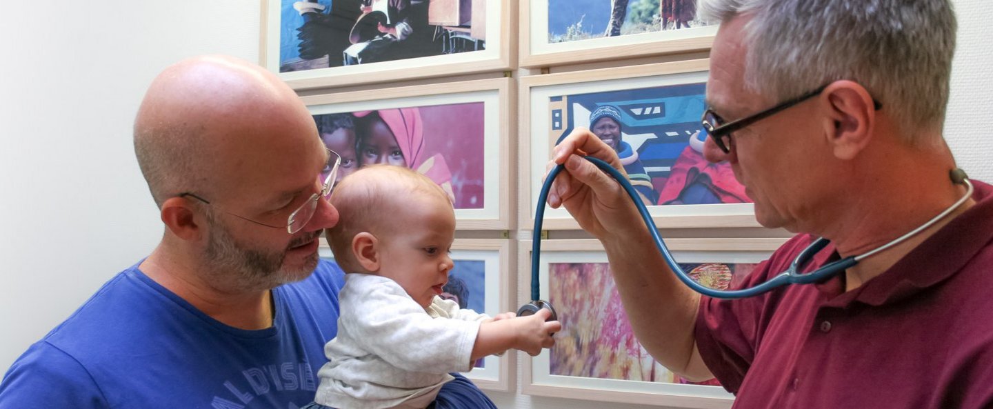 Mann hält Baby auf dem Arm, das nach dem Stethoskop des gegenüber stehenden Arztes greift.