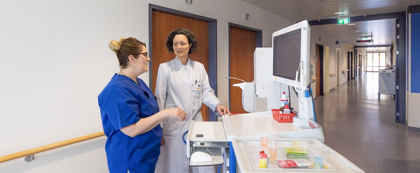 Ärztin und Pflegefachfrau im Gespräch am Patientenwagen.