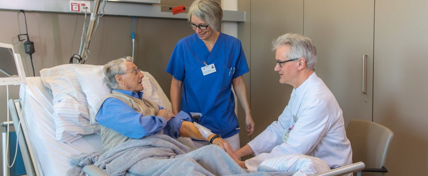 Arzt und Pflegefachfrau im Gespräch mit Patient, der im Klinikbett liegt und ein Pflaster am Arm hat.