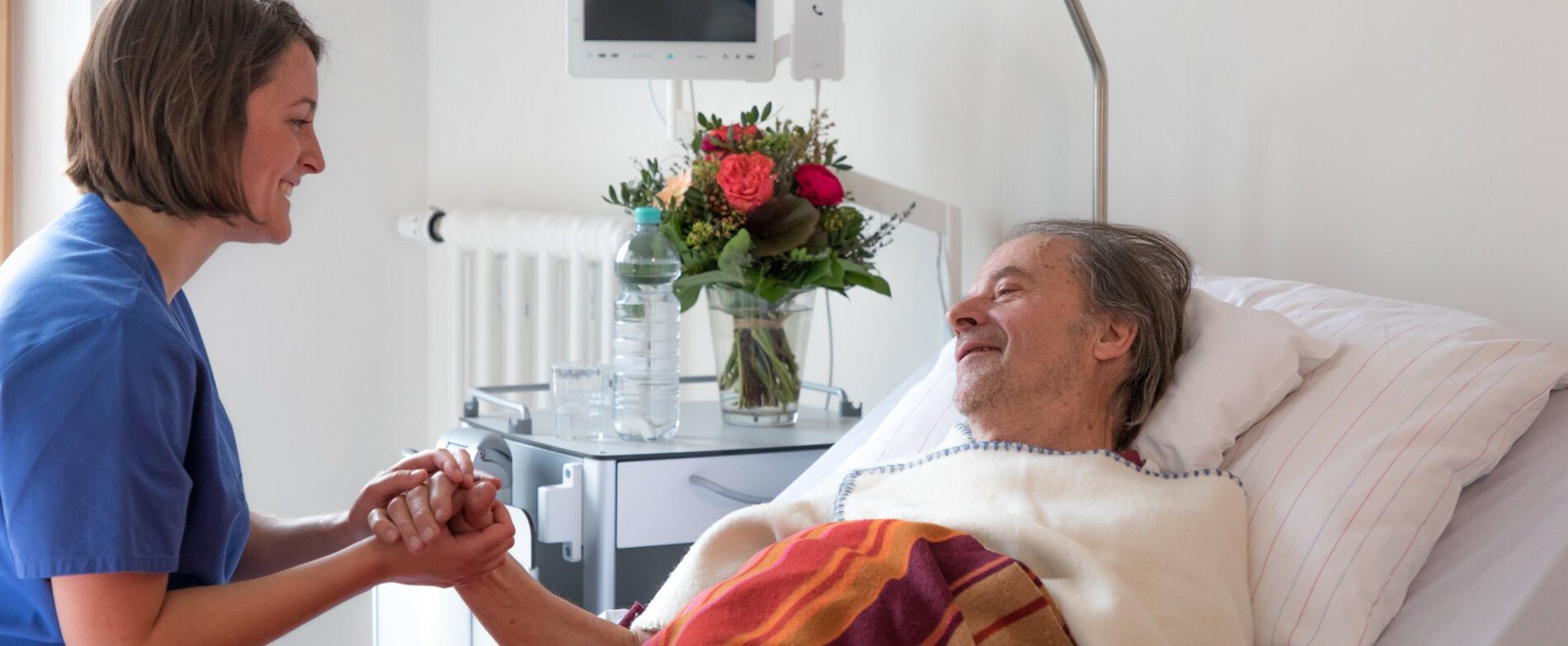 Pflegefachfrau sitzt am Bett des Patienten und hält seine Hand, während sie sich unterhalten.