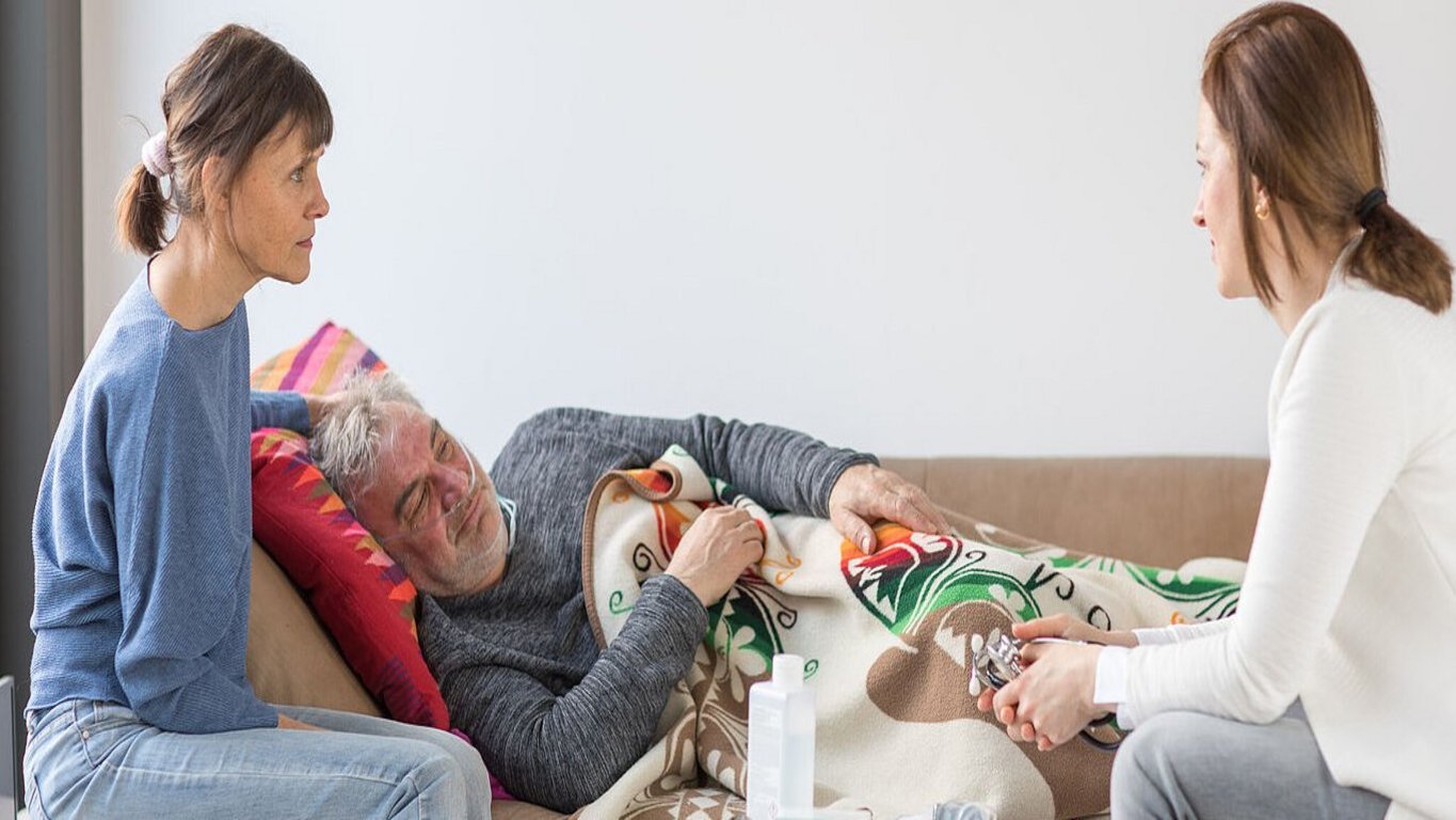 Ärztin im Gespräch mit liegendem Patienten und Angehöriger in häuslicher Umgebung.